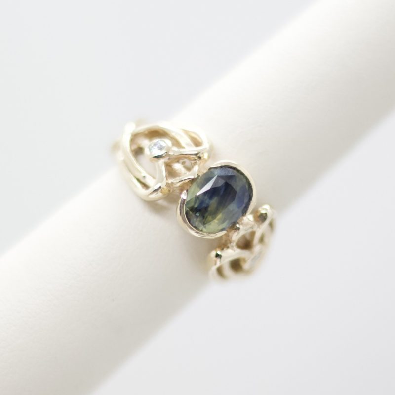 This ring is unique with its bi-colored sapphire center stone, two accent diamonds, and the lattice work of the band in 14K yellow gold. This would be a stunning statement ring for anyone. Designed by Morgan's Treasure Sapphire: Natural oval, bi-colored (blue, green, and yellow) sapphire, 8mm x 6mm, 2.16ct Diamonds: 0.06ctw Dimensions: 10.5mm W Free ring sizing with purchase -- contact us to select your finger size
